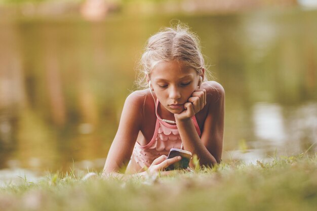 Cute little girl enjoying summer, looking at the smartphone, relaxing and daydreaming near the lake fringed with lush scenery.