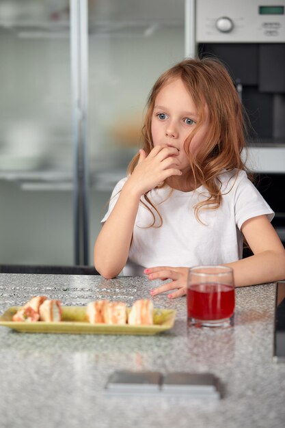 Cute little girl enjoys eating sushi Cheerful funny caucasian blond preschool child girl with sushi roll and On kitchen background