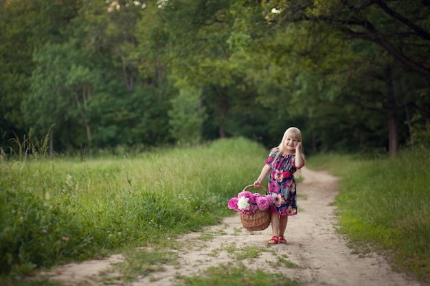 Cute little girl on a forest road with a basket of flowers The concept of carefree childhood