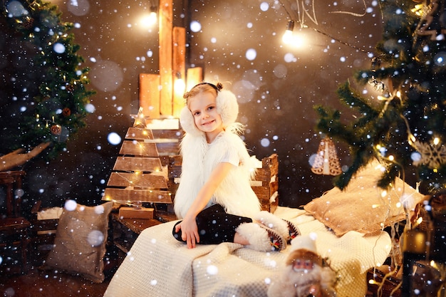 Cute little girl in fur earmuffs sitting near Christmas tree