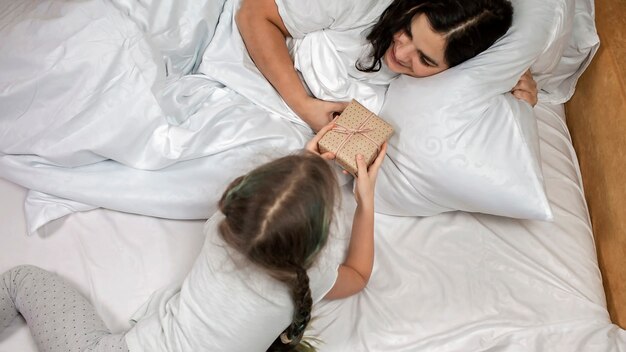 Cute little girl giving surprise and gift to her mom in the bed