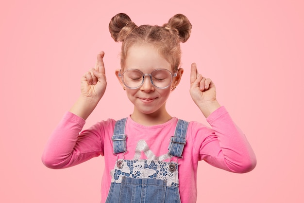 Bambina sveglia in occhiali e abbigliamento casual con gli occhi chiusi e le dita incrociate esprimendo desiderio stando in piedi