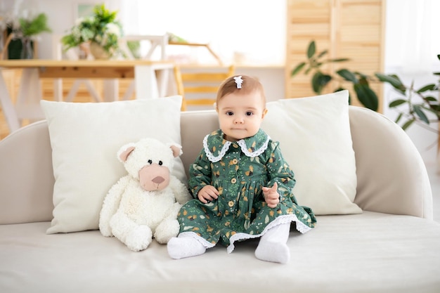 A cute little girl in a green cotton dress is sitting on the sofa with a white teddy bear in the living room of the house looking at the camera smiling the baby is at home