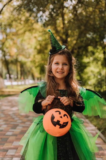 お菓子のカボチャのバスケットを持つ魔女や妖精の緑のハロウィーンの衣装でかわいい女の子