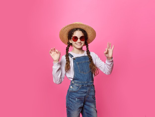 Cute little girl in hat and red sunglasses looking at camera showing a ok sigh isolated pink background Front portrait