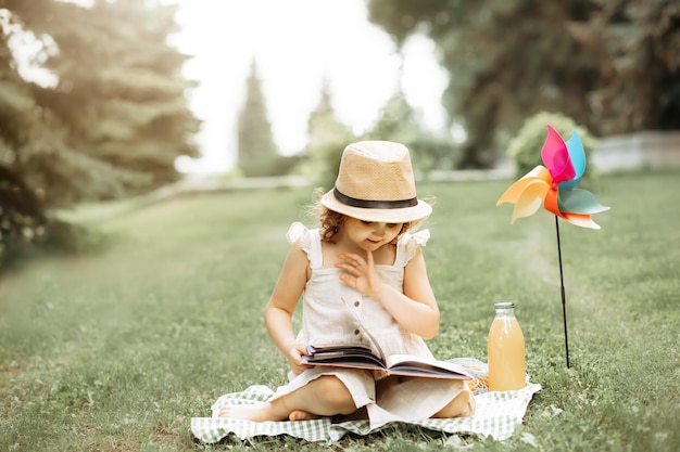 Cute little girl in a hat sitting on a blanket and reads a book at a picnic in nature