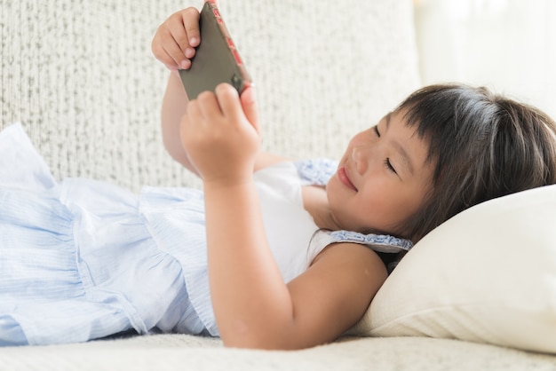 Cute little girl having fun to play game on smart phone lying on sofa