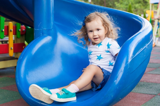 Bambina sveglia divertendosi in un campo da giuoco all'aperto un giorno di estate soleggiato. bambino su scivolo di plastica. attività divertente per bambini.