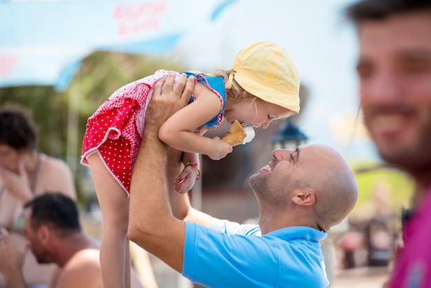 cute little girl having fun with her young father while eating ice cream by the sea during summer vacation holidays, celebration, children and people concept