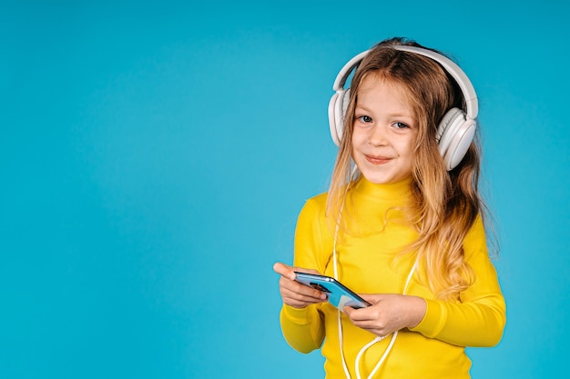 Cute little girl in headphones is using a smartphone, looking at camera and smiling isolated