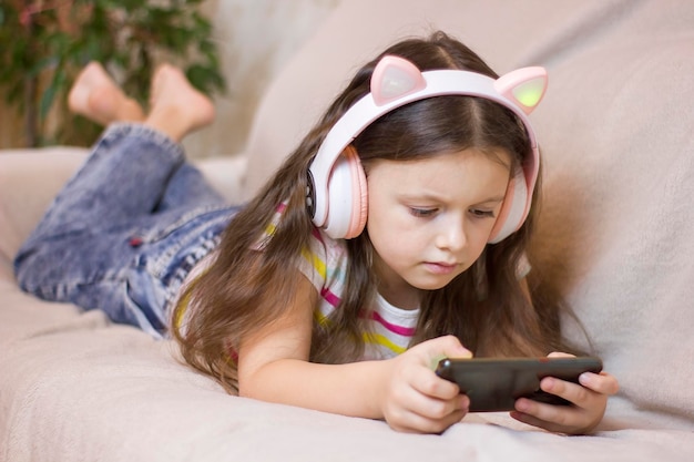 Photo cute little girl in headphones is using a smartphone smiling while lying on the sofa at home