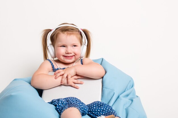 Cute little girl in headphones listening to music using a tablet and smiling while sitting on blue b...