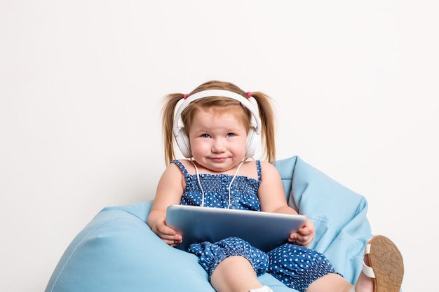 Милая маленькая девочка в наушниках слушает музыку с помощью планшета и улыбается, сидя на синем б ...
