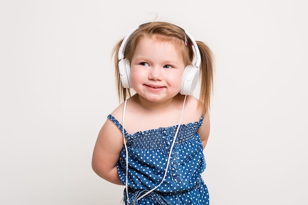 Милая маленькая девочка в наушниках слушает музыку с помощью планшета и улыбается на белом фоне