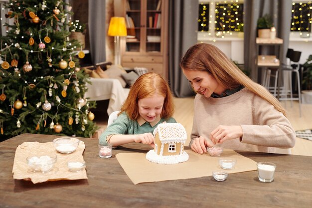 Cute little girl helping her mother sprinkle roof of tasty gingerbread house decorated