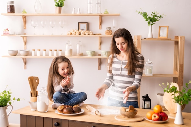 かわいい女の子と彼女の美しい母親が小麦粉を生地に振りかけ、焼きながら笑顔