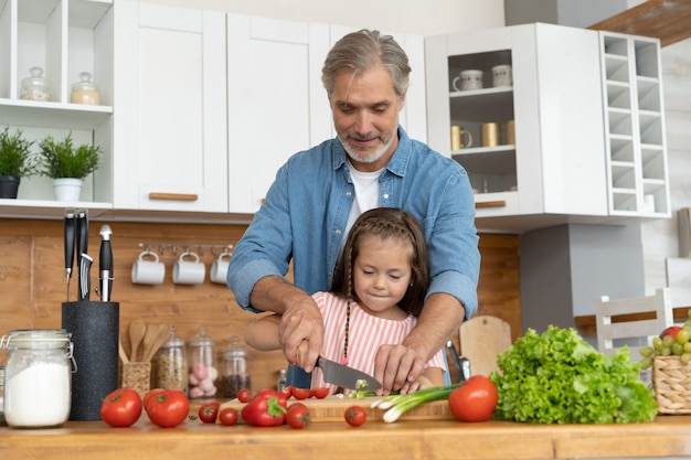 Милая маленькая девочка и ее красивый папа режут овощи и улыбаются, готовя дома на кухне.