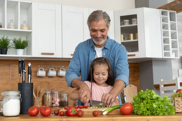 Cute little girl and her handsome dad are cutting vegetables and smiling while cooking in kitchen at home.