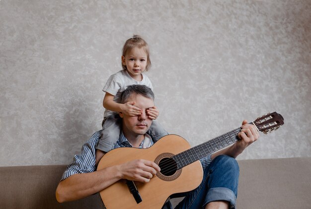 Photo cute little girl and her handsome father play guitar and smile sitting on the couch at home. father's day. care and education of children.