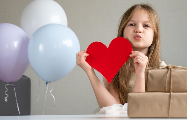 Cute little girl holding big red paper heart and blows a kiss congratulating mom on Mother's day
