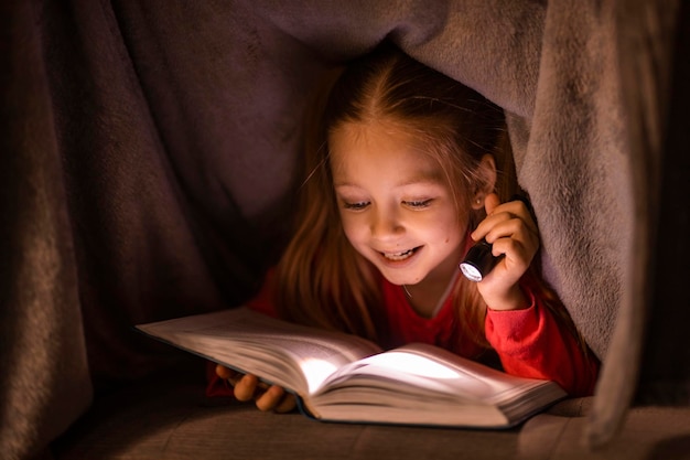 Cute Little Girl Holding Flashlight And Reading Book Under Blanket At Home