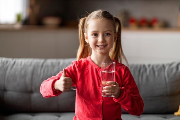 Cute little girl holding glass of water and showing thumb up