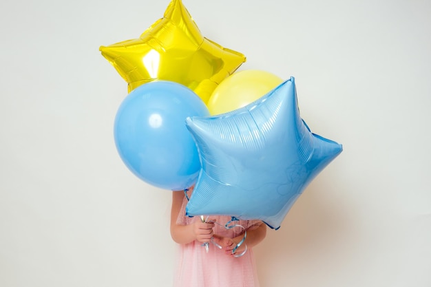 Cute little girl holding helium blue, yellow and gold balloons on white background in studio