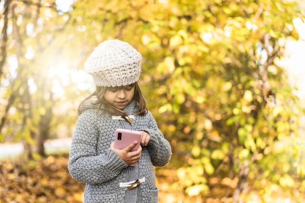 Cute little girl holding smartphone at park during foliage time Little girl using a phone typing and taking photos of yellow autumnal landscape Childhood and activist concept for the planet Earth