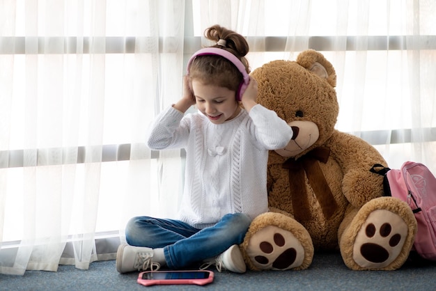 Cute little girl holding tablet computer in her hands. She is in headphones. Advertising new gadget, educational app. She is near the window. Games. Children use technology.