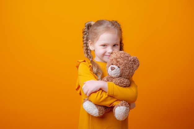 Cute little girl holding a teddy bear on a yellow background hugging