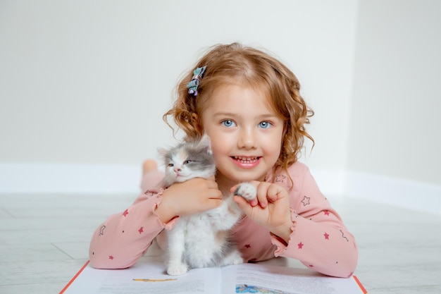 Una bambina carina abbraccia un gattino sdraiato sul pavimento della casa che legge un libro