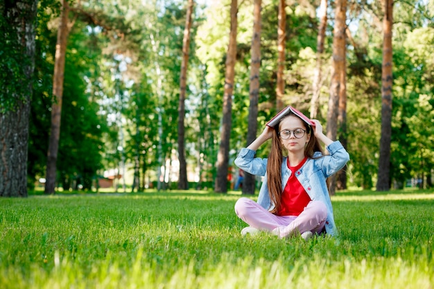 Фото Милая маленькая девочка в очках с книгой на голове в летнем парке