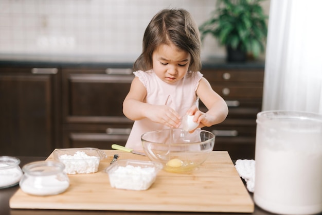 Cute little girl is breaking an egg into a mixing bowl at the kitchen. Child is learning to bake cake standing breaking eggs into a bowl at kitchen.