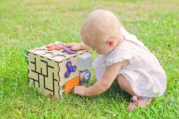 Cute little girl is playing with busiboard outdoors on green grass. educational toy for toddlers. girl opened door to cube of board.