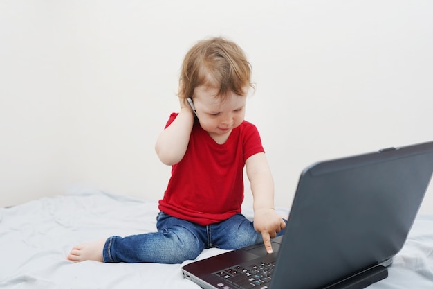 Cute little girl is sitting on bed with her laptop and phone holding near ear