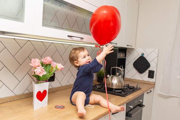 A cute little girl is sitting in the kitchen and playing with a red heartshaped ball