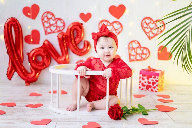 a cute little girl is sitting in a red bodysuit against a background of red hearts and the inscription love. the concept of valentine's day, valentine's day