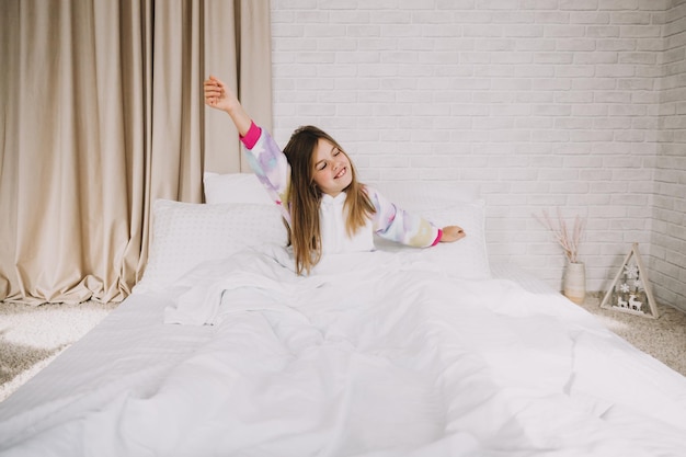 A cute little girl is stretching in bed in her bedroom on white linens.