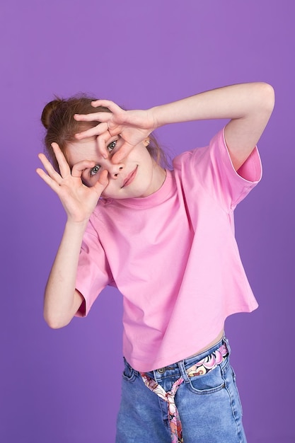 A cute little girl in jeans and a pink Tshirt on a purple background in the studio smiling grimacing and looking at the camera Children's games