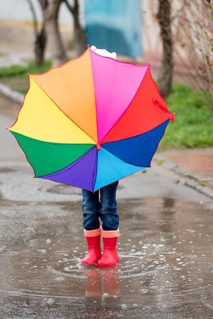 かわいい女の子が水たまりに飛び込んで楽しんでいる女の子は彼女の手に虹の傘を持っています