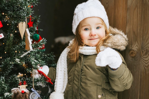 cute little girl in knitted white hat stand at porch of country house and decorate Christmas tree fo