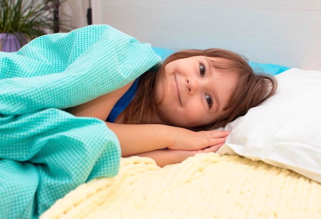 A cute little girl laughs as she lies in bed under blue wool blanket. Happy morning.