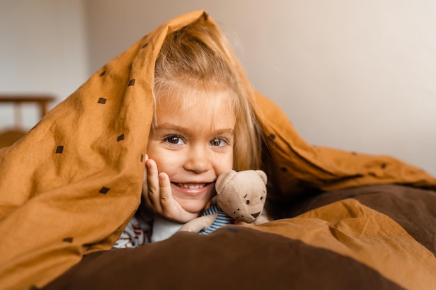 Cute little girl lies cozy in bed covered with a blanket over her head smiling with copy space
