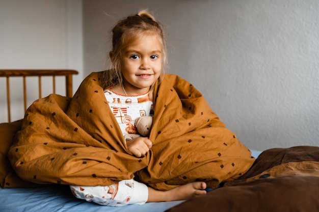 Cute little girl lies cozy in bed covered with a blanket over her head smiling with copy space