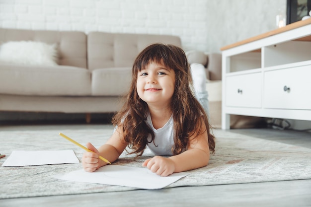 Una bambina carina giace sul pavimento della casa e disegna con le matite il tempo libero dei bambini