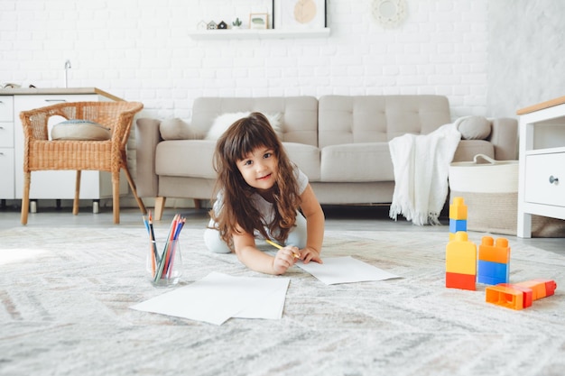 A cute little girl lies on the floor of the house and draws with pencils children's leisure