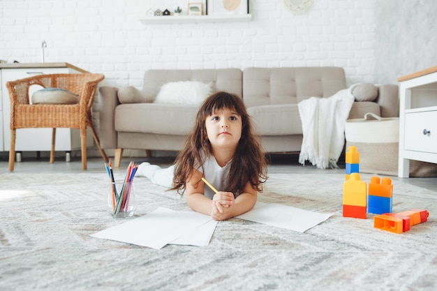 A cute little girl lies on the floor of the house and draws with pencils children's leisure