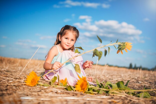 Cute little girl in a long sundress and with sunflowers sits on a field with mowed wheat on a sunny warm summer day