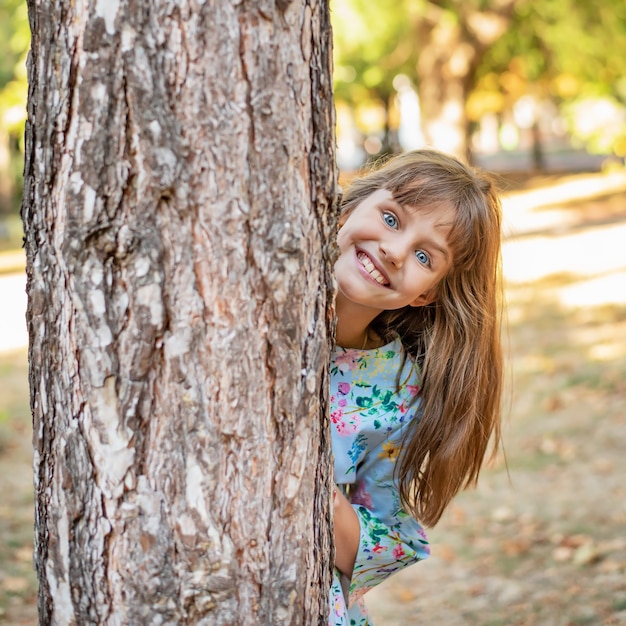 Foto una bambina carina guarda fuori da dietro un albero nel parco con un sorriso.