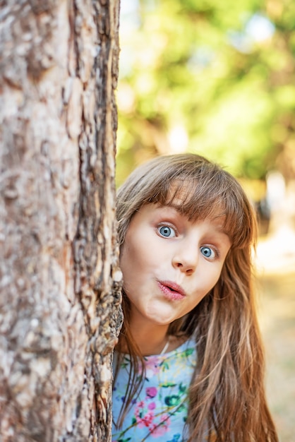 公園の木の後ろからかわいい女の子が外を眺めています。